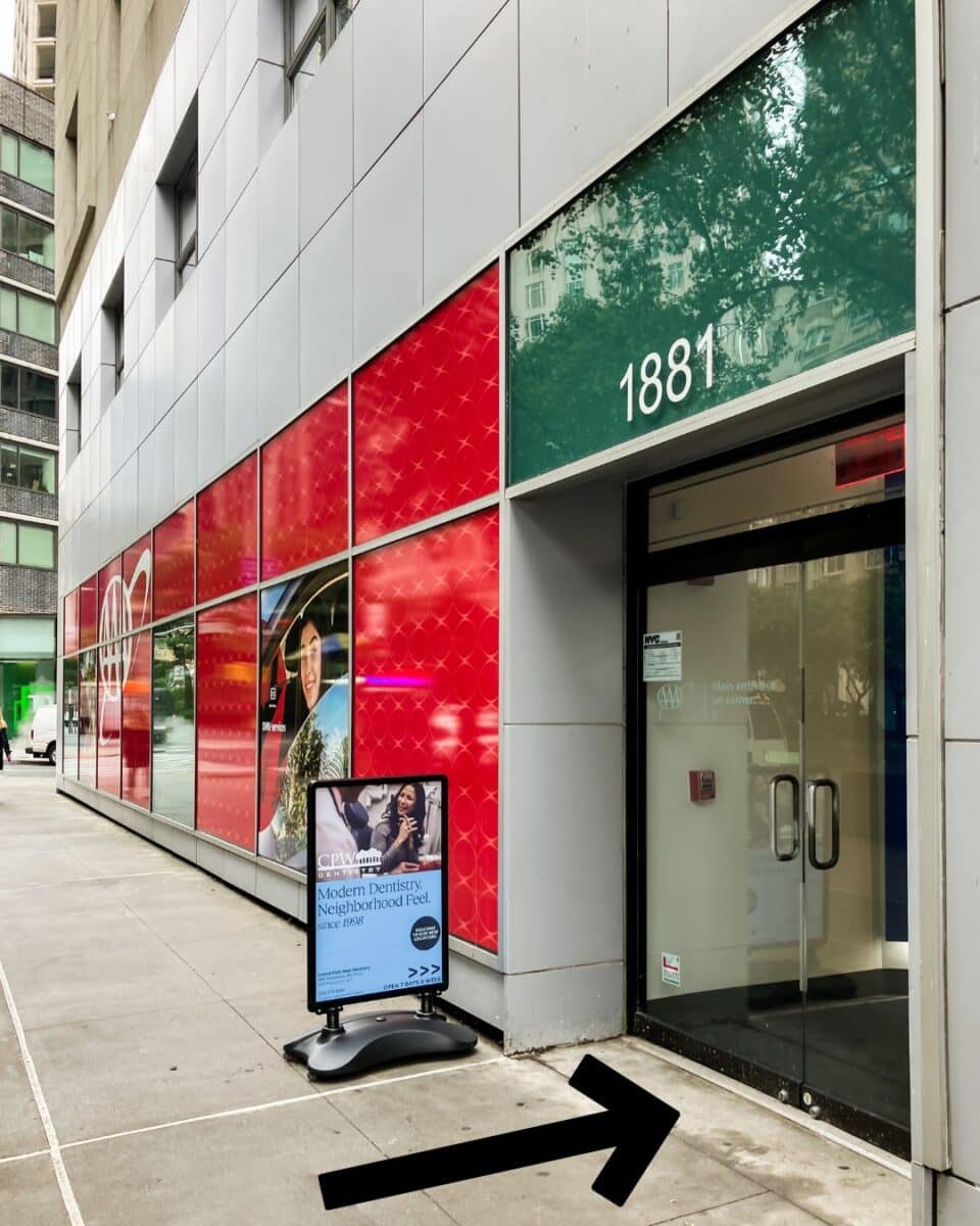 Exterior view of 1881 Broadway with an arrow pointing toward the correct entrance for CPW Dentistry, located between 62nd and 63rd Streets. A sidewalk sign directs visitors to the Broadway entrance rather than the AAA office at the corner.