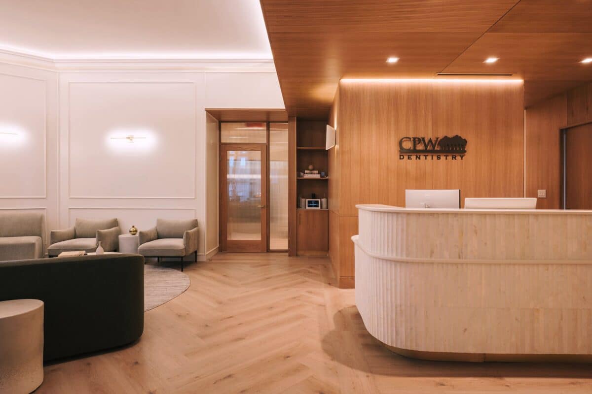 A contemporary office waiting area with neutral-toned armchairs, light wood furniture, and soft lighting. The CPW Dentistry logo is visible behind the reception desk.