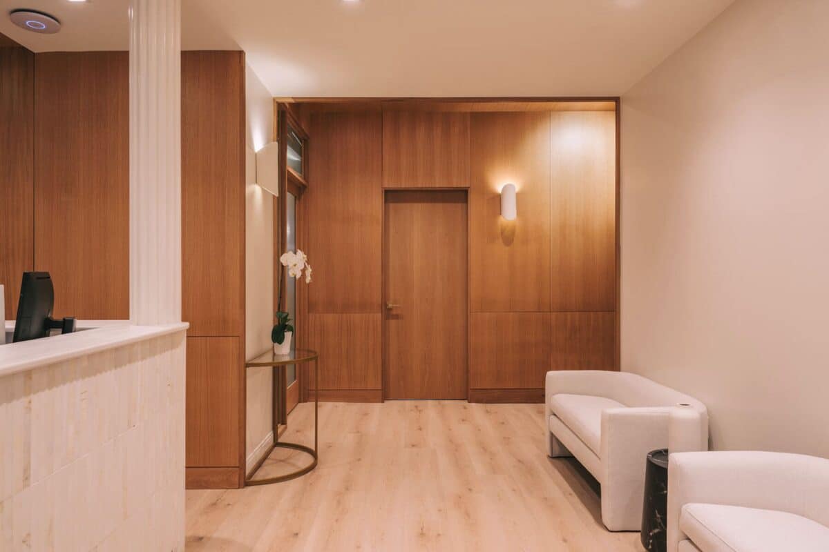 Reception area with warm wood-paneled walls, a closed wooden door, light wood flooring, and cream-colored lounge chairs.